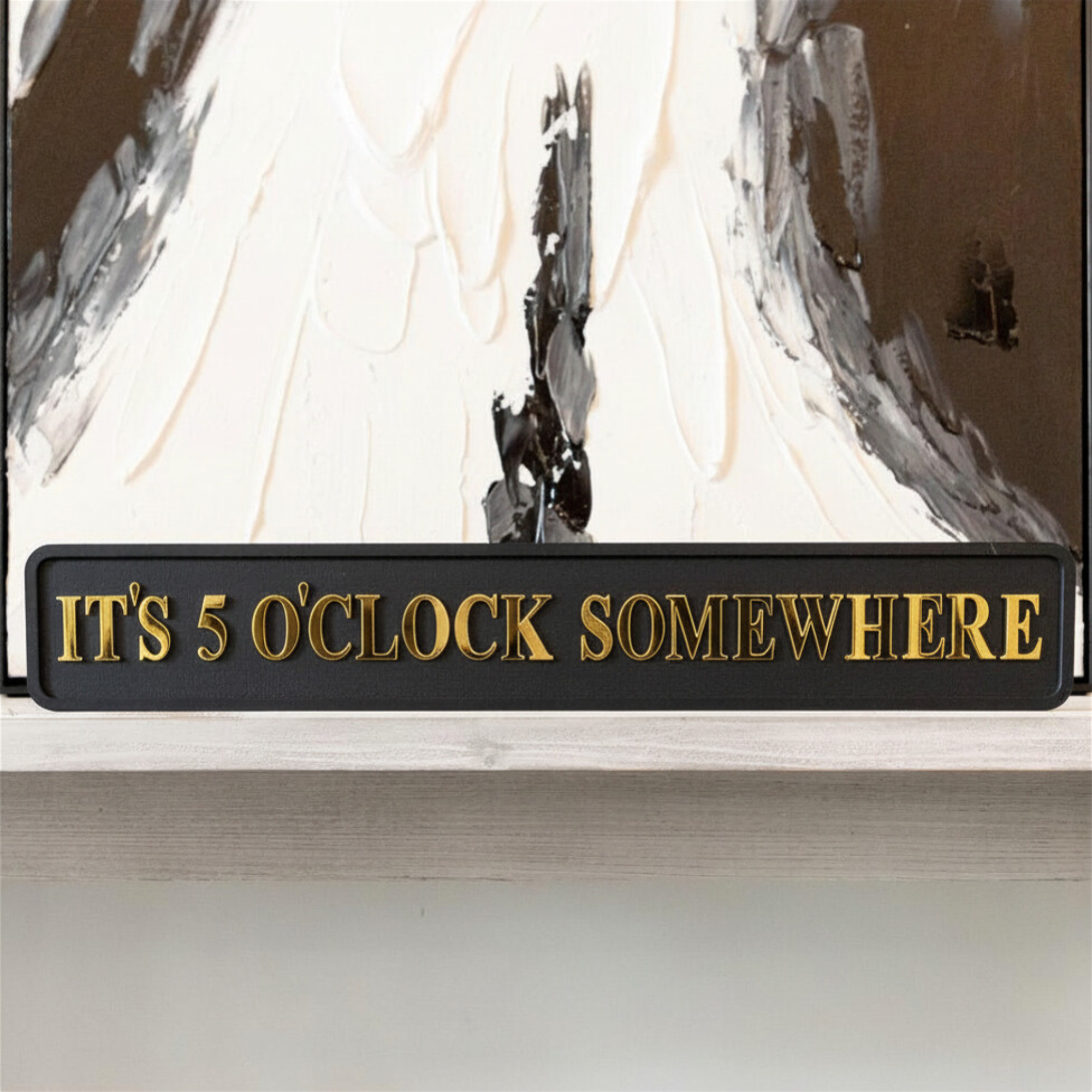 Decorative sign with 'It's 5 o'clock somewhere' text in front of abstract art