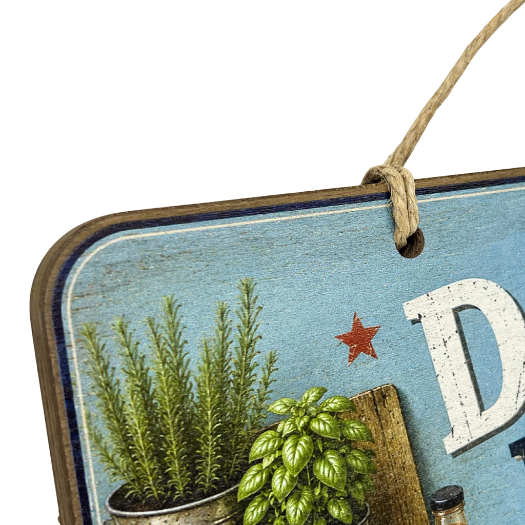 A rustic decorative hanging sign with a distressed light blue background, a brown wooden border, a hanging twine loop, and printed botanical illustrations including green plants and a small red star. Part of a large white letter and a bottle illustration are visible.