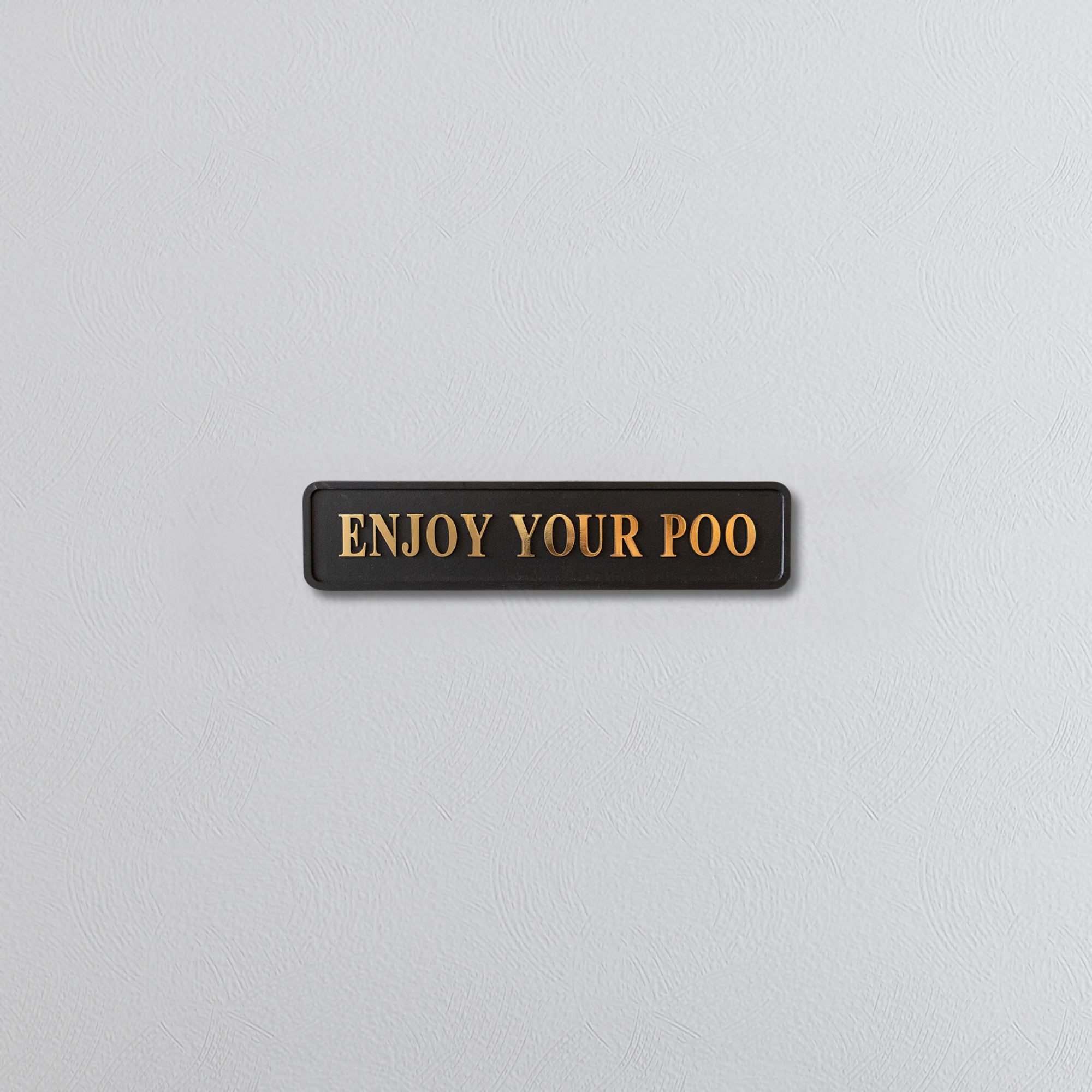 Black sign with gold text 'ENJOY YOUR POO' on a light gray background