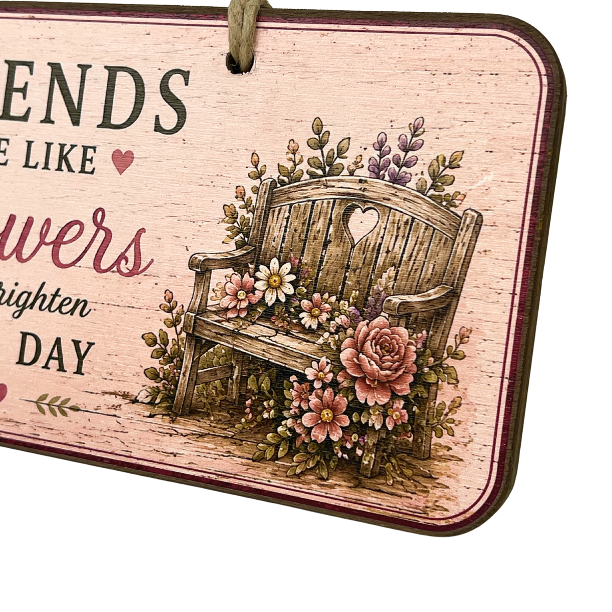 A rectangular decorative wall sign with rounded corners, hanging by a rope loop. The sign has a distressed light pink wood-grain background with printed text and an illustration of a wooden bench surrounded by pink and white flowers and green leaves.