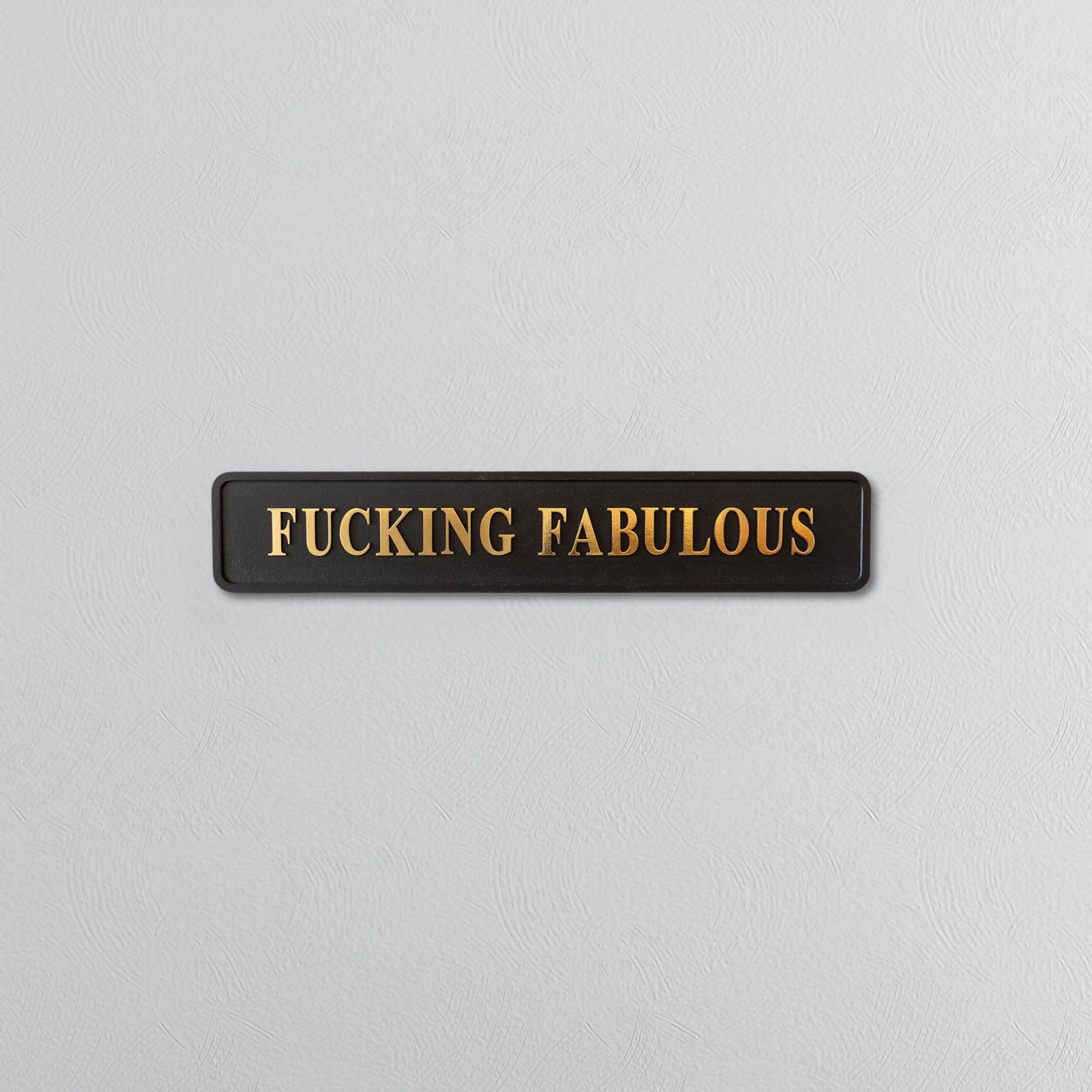 Black rectangular sign with 'FUCKING FABULOUS' in gold letters on a light gray background