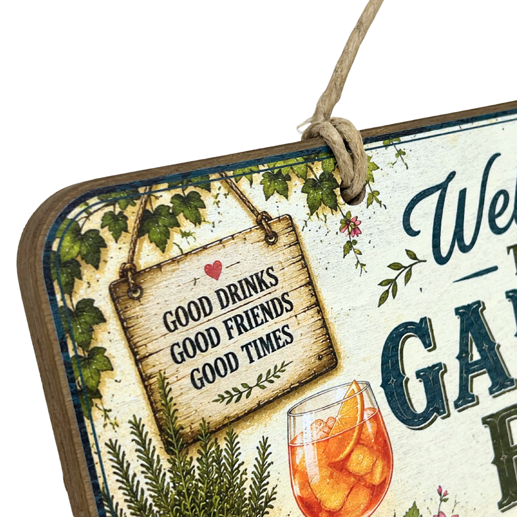 A decorative hanging sign with a rustic wood-look frame and twine loop, featuring the phrase “GOOD DRINKS GOOD FRIENDS GOOD TIMES” on a small wooden plaque. The sign includes green ivy, a glass of iced tea with orange slices, and partial decorative text on a cream background.