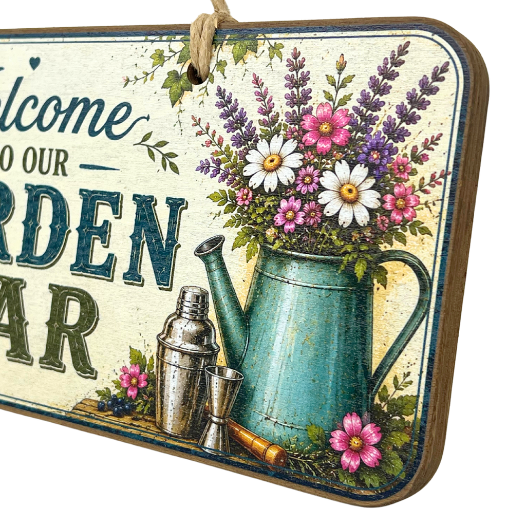 A rectangular hanging garden sign with a rustic distressed cream background and a wooden border. The sign features vintage-style text reading 'Welcome to our GARDEN BAR' alongside an illustration of a teal watering can filled with pink, purple, white, and yellow flowers, plus a metal shaker and small jugs.
