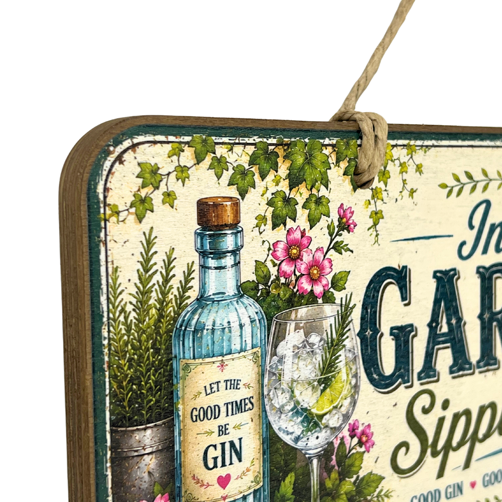 A decorative rectangular hanging sign with a rustic wood edge and twine loop, featuring an illustrated blue gin bottle, a filled cocktail glass, pink flowers, green ivy, and partial text reading 'In A GIN' and 'Good Times Be Gin.'.