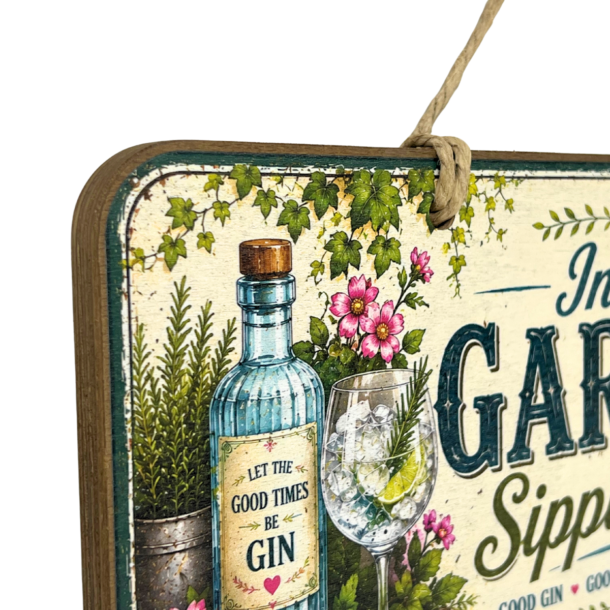 A decorative rectangular hanging sign with a rustic wood edge and twine loop, featuring an illustrated blue gin bottle, a filled cocktail glass, pink flowers, green ivy, and partial text reading 'In A GIN' and 'Good Times Be Gin.'.