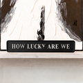 Black decorative sign with HOW LUCKY ARE WE silver text in front of an abstract painting