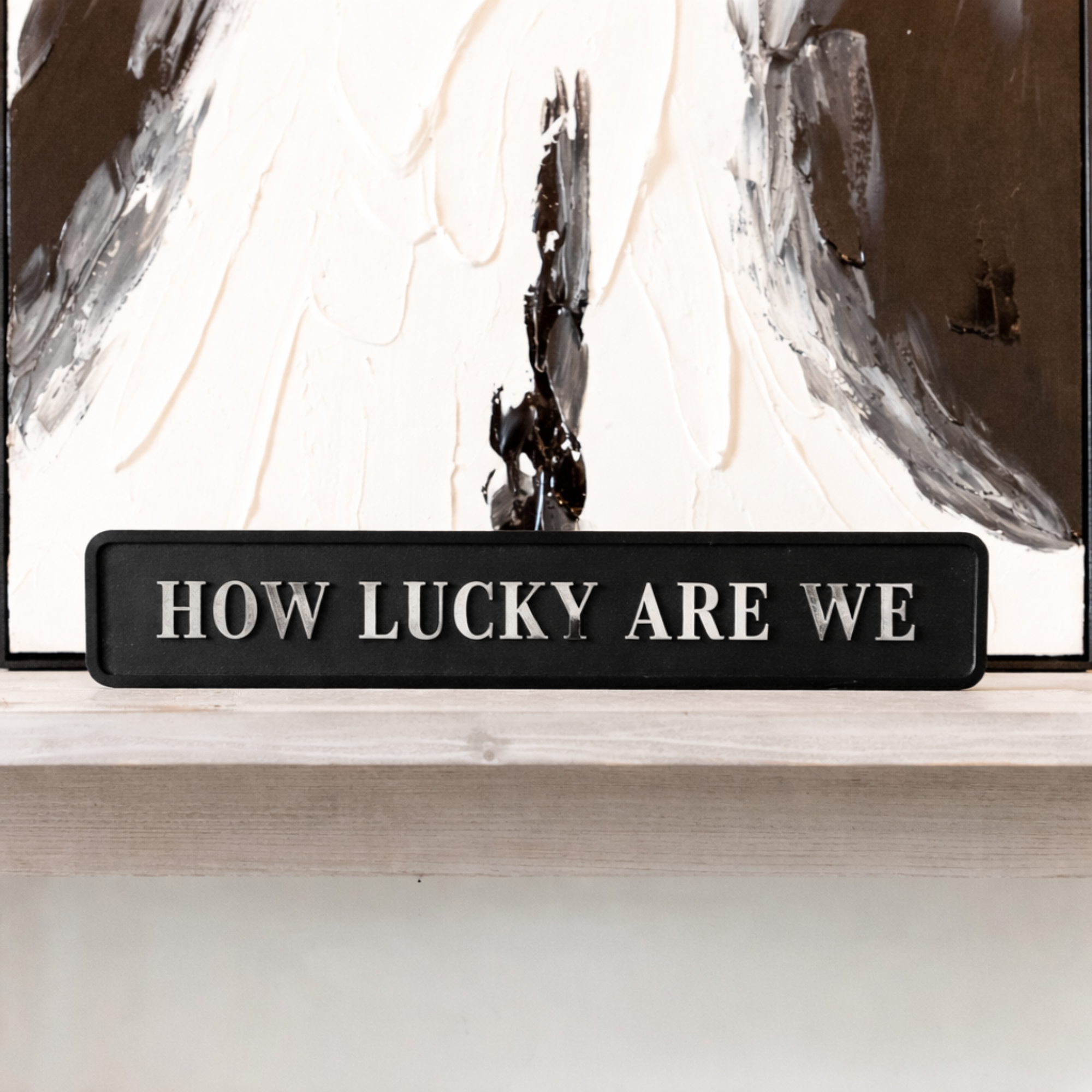 Black decorative sign with HOW LUCKY ARE WE silver text in front of an abstract painting