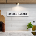 Kitchen wall with 'HUSTLE HARDER' sign, white tiles, wooden cabinets, and decor items.