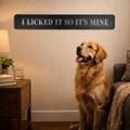 Dog sitting in a living room with a sign above it that reads 'I licked it so it's mine'.