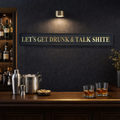 Bar setup with drinks and a humorous sign 'LETS GET DRUNK & TALK SHITE' on a dark wall.