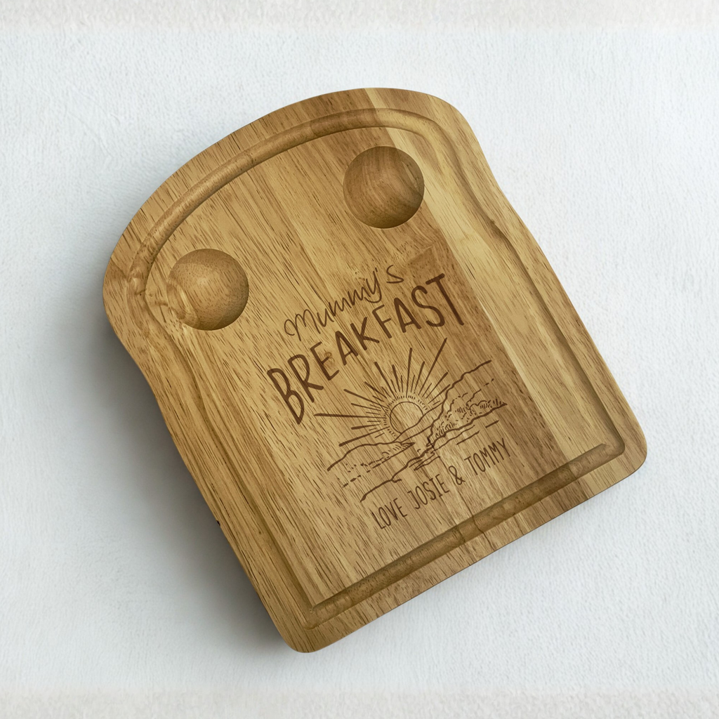 An engraved wooden toast-shaped board with 2 carved pockets at the top for holding eggs, an engraved sunset design with 'mummy's breakfast' above and a personalised message at the bottom.