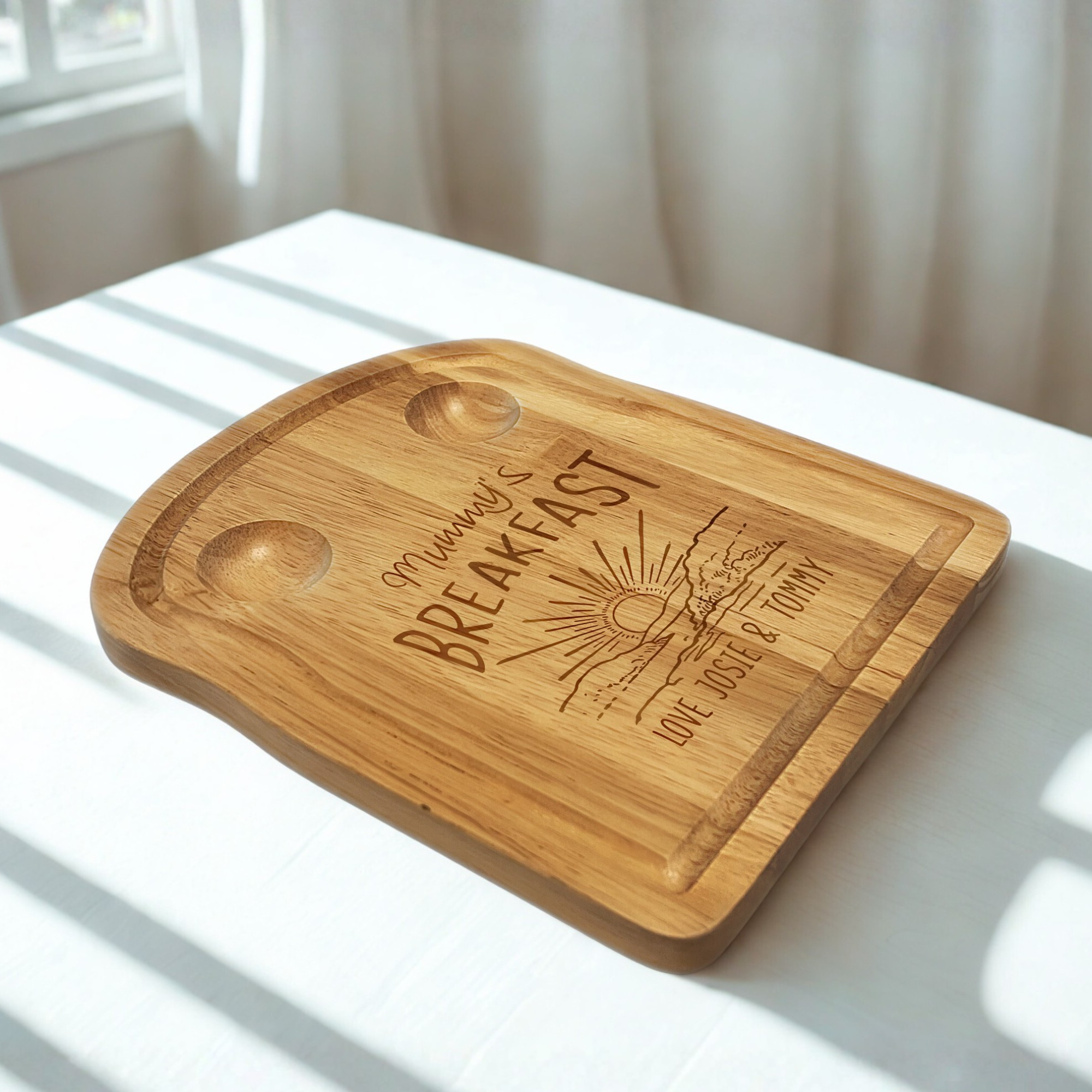 An engraved wooden toast-shaped board with 2 carved pockets at the top for holding eggs, an engraved sunset design with 'mummy's breakfast' above and a personalised message at the bottom. On a white table in the sunshine.