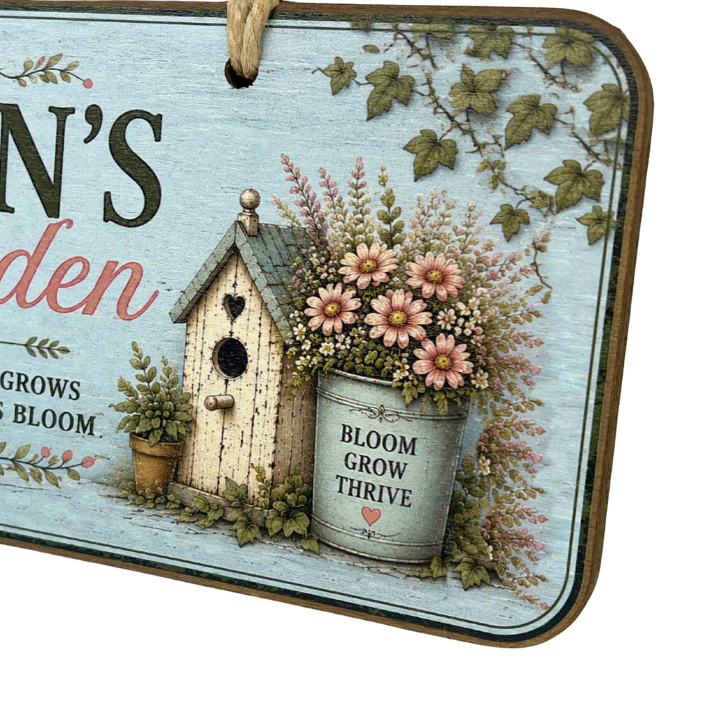 A decorative rectangular garden sign with a rustic light blue background, featuring a small white birdhouse, a metal bucket planter filled with pink flowers and greenery, and partial text reading 'Garden' and 'Bloom Grow Thrive.'.
