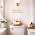 Cream and gold decorative sign with the words 'NICE ASS' displayed in a stylish bathroom