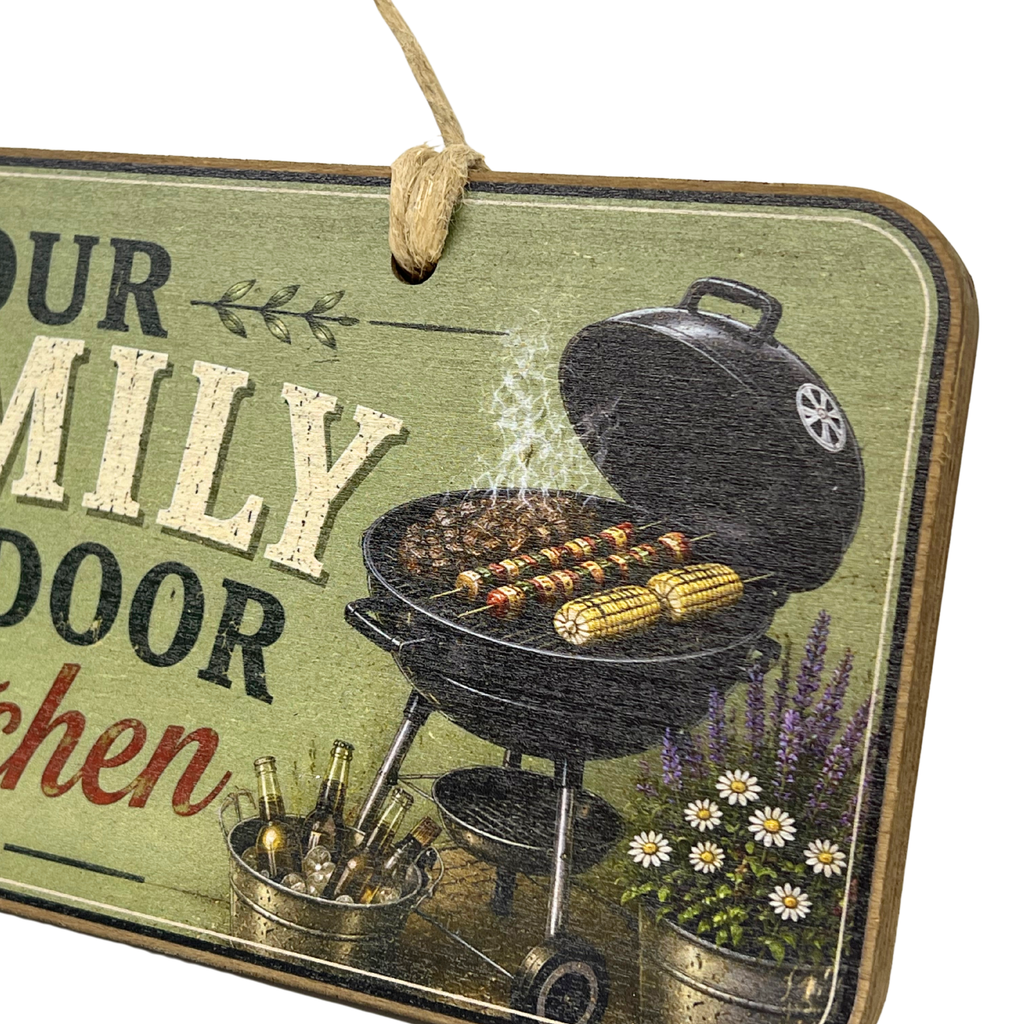 A rectangular hanging wooden sign with a rustic distressed finish, featuring an illustration of a charcoal grill with corn on a grate, cooking smoke, bottles in a bucket, and white daisies in a pot. The sign has a green background with text that reads 'OUR FAMILY DOOR kitchen' and is suspended by a twine loop.