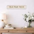 Decorative motivational wall sign reading 'PRAY WAIT TRUST' with gold text above a wooden console table with lamp, books, and flowers.