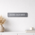 Grey sign with 'SAME OLD SHIT' silver text on a white wall with decor items.