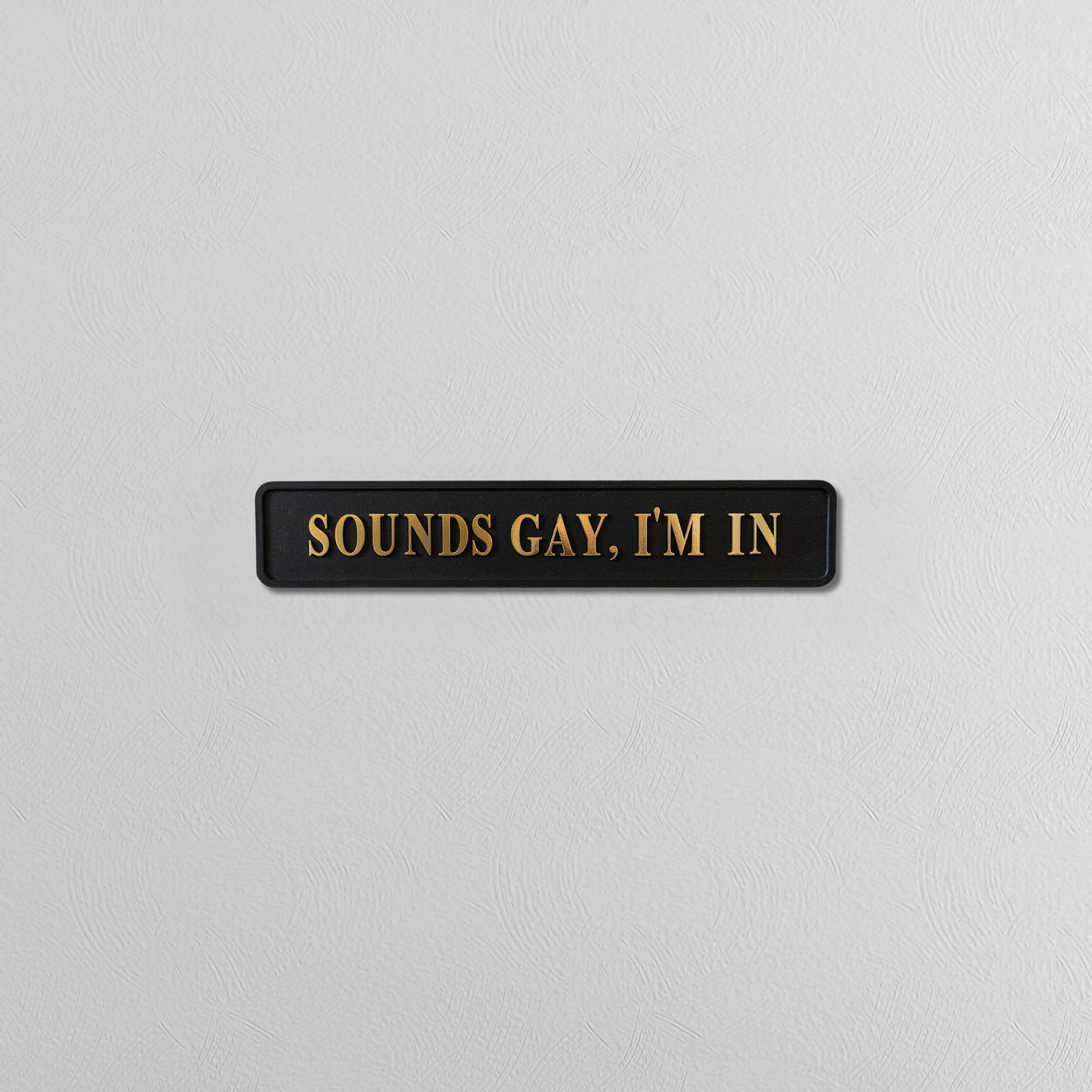 Black rectangular sign with gold text 'SOUNDS GAY. I'M IN' on a light gray background