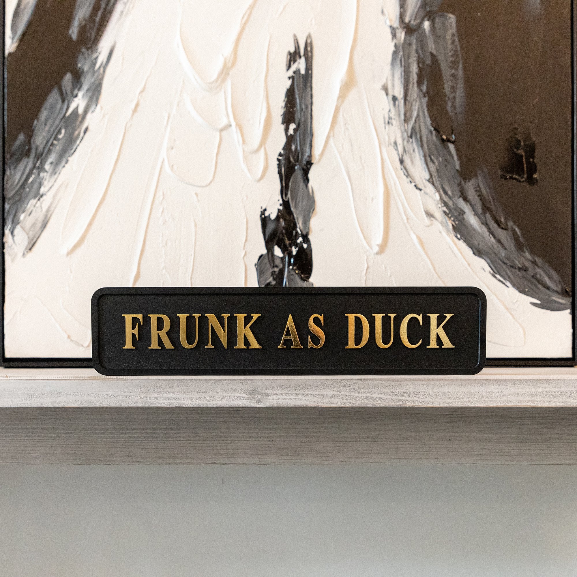 Decorative sign with 'FRUNK AS DUCK' gold foiled text in front of a abstract painting.