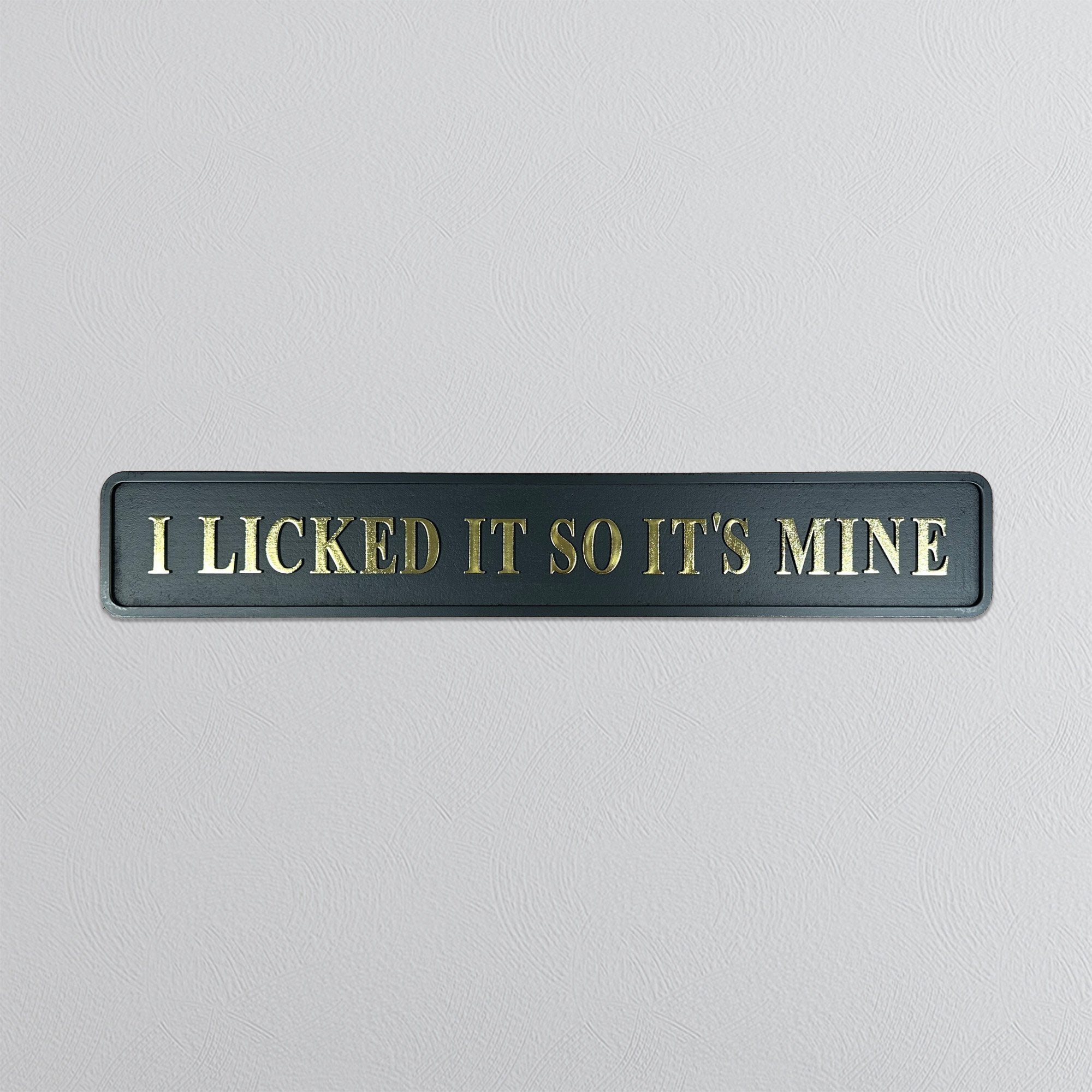 Black sign with gold text 'I LICKED IT SO IT'S MINE' on a light gray background