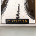 Decorative sign with 'IT IS WHAT IT IS' text in front of an abstract painting.