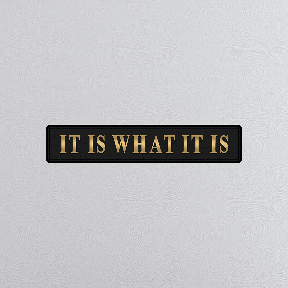 Black rectangular sign with gold text 'IT IS WHAT IT IS' on a light gray background