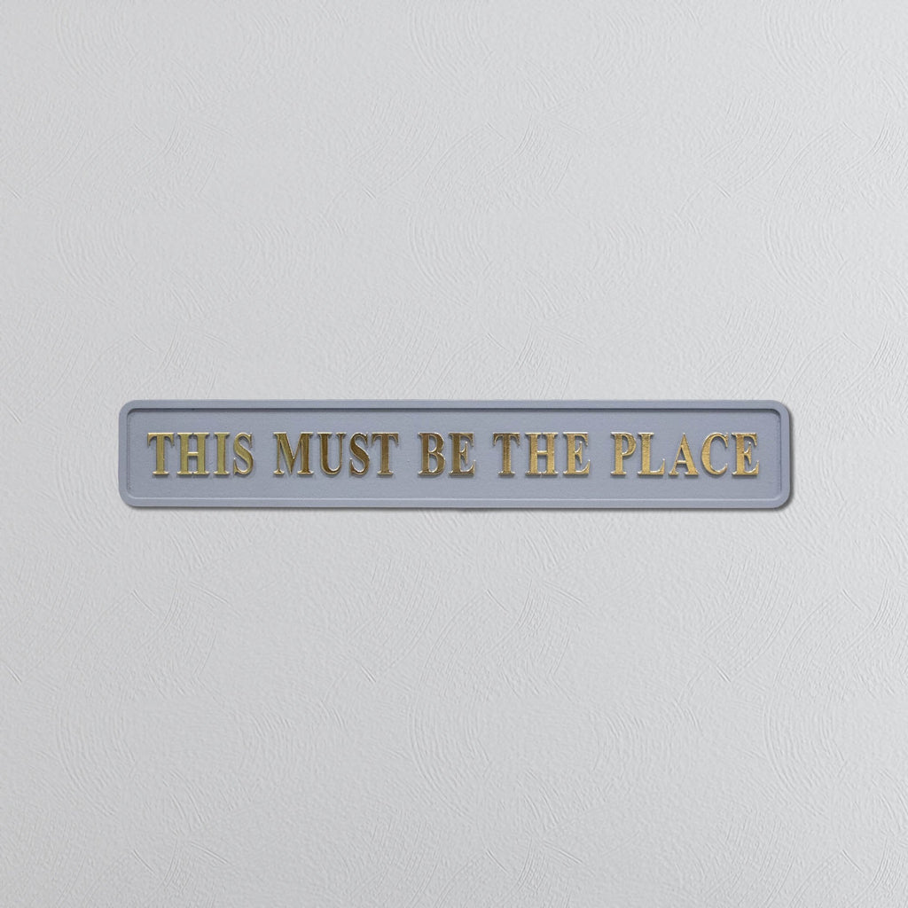 Decorative grey sign with 'THIS MUST BE THE PLACE' in gold text on a light gray background