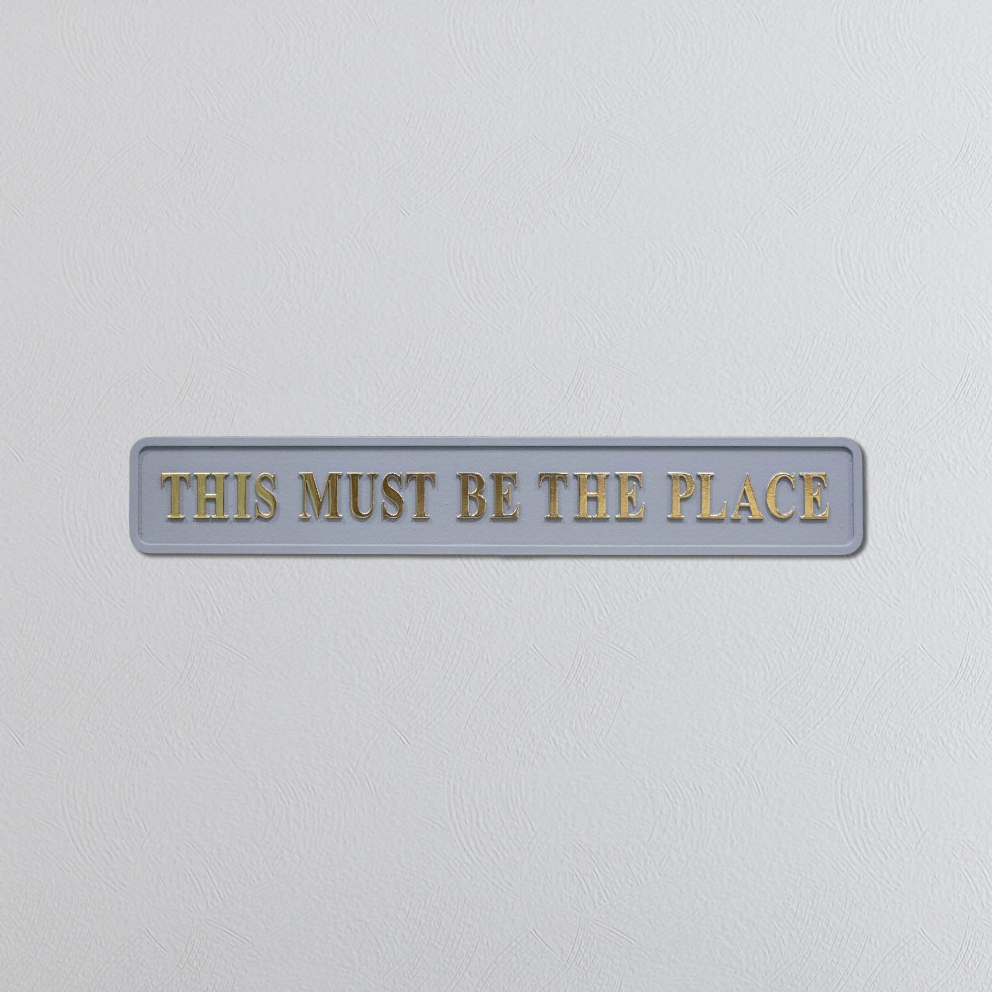 Decorative grey sign with 'THIS MUST BE THE PLACE' in gold text on a light gray background