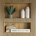 Wooden shelf with decorative items including a plant, vases, and a cream sign reading NOBODY GETS OUT SOBER in gold text.