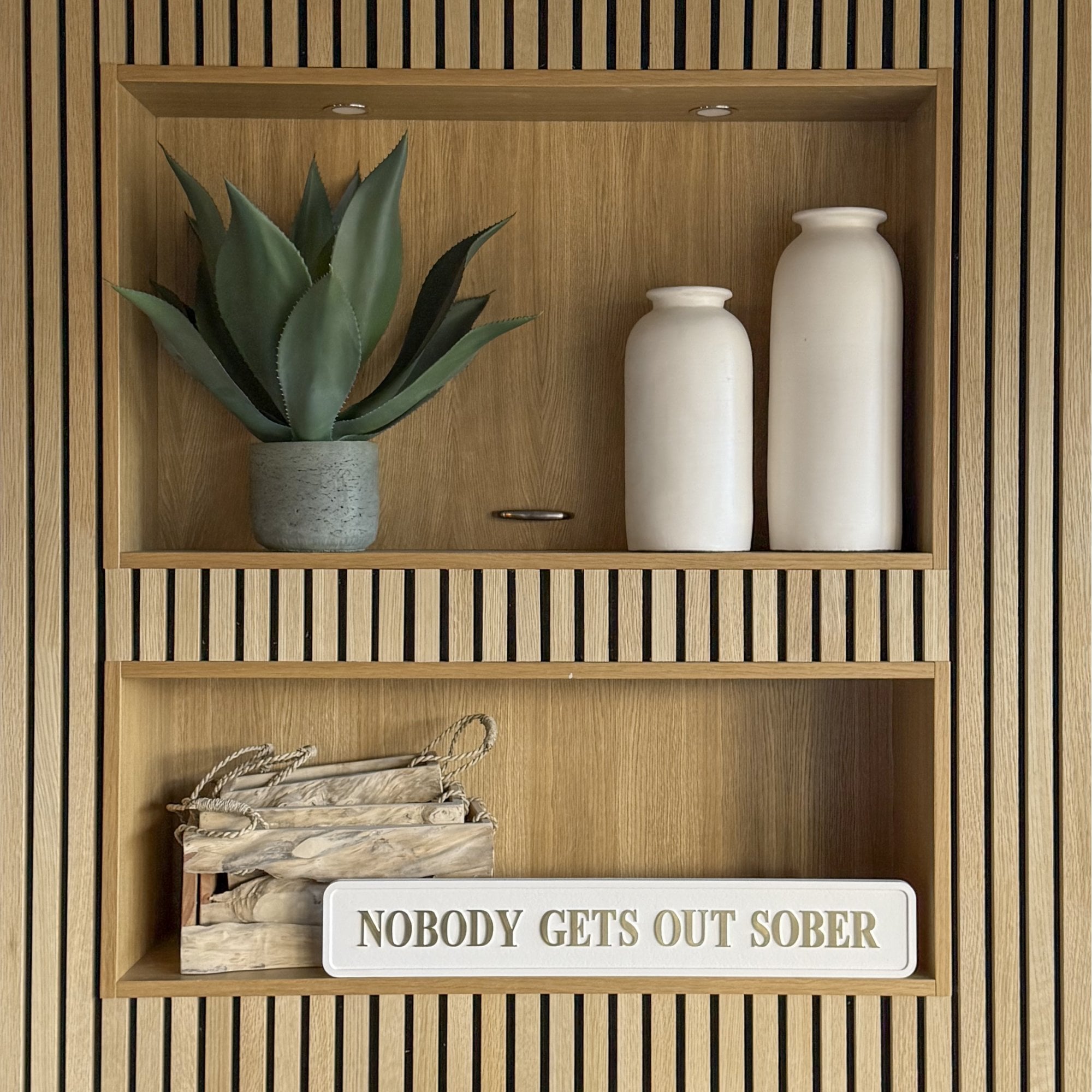Wooden shelf with decorative items including a plant, vases, and a cream sign reading NOBODY GETS OUT SOBER in gold text.