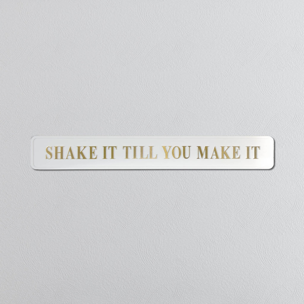 Rectangular white sign with 'SHAKE IT TILL YOU MAKE IT' in gold text on a light gray background