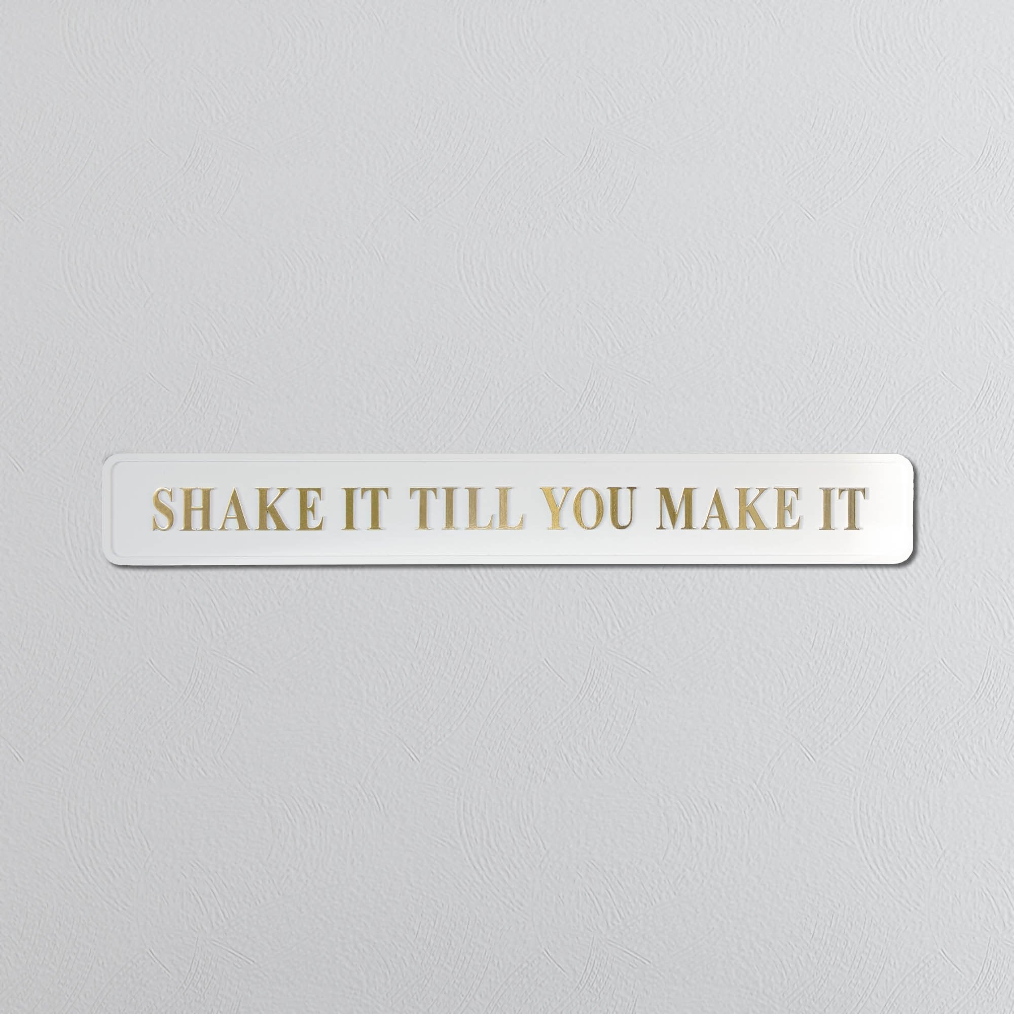 Rectangular white sign with 'SHAKE IT TILL YOU MAKE IT' in gold text on a light gray background