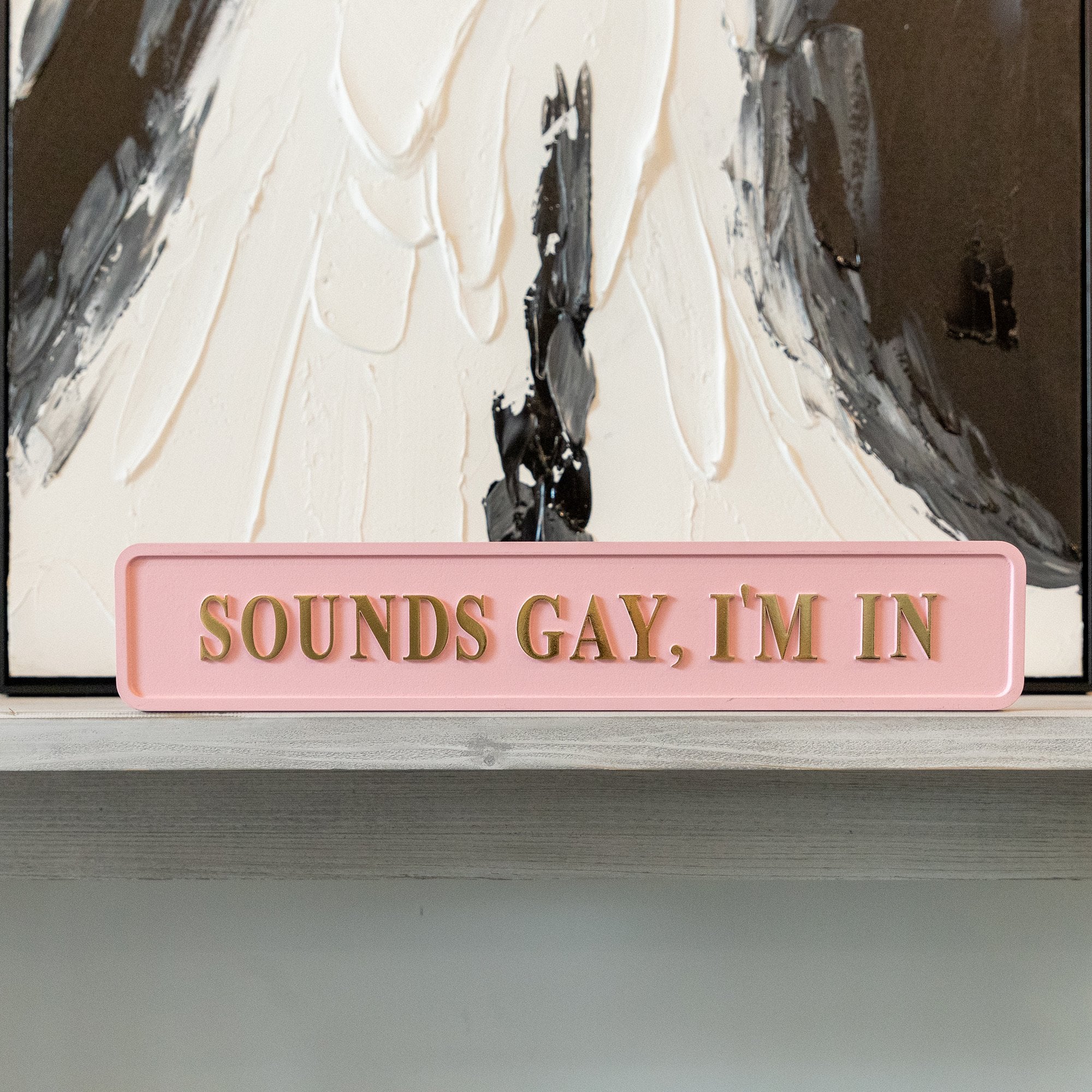 Pink sign with gold text 'SOUNDS GAY, I'M IN' in front of a painting