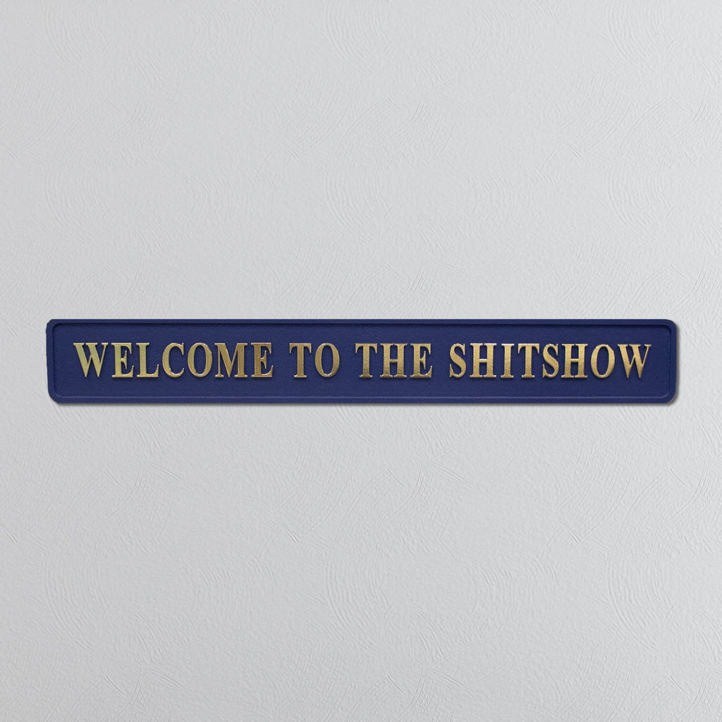 Navy blue sign with gold text 'WELCOME TO THE SHITSHOW' on a light gray background