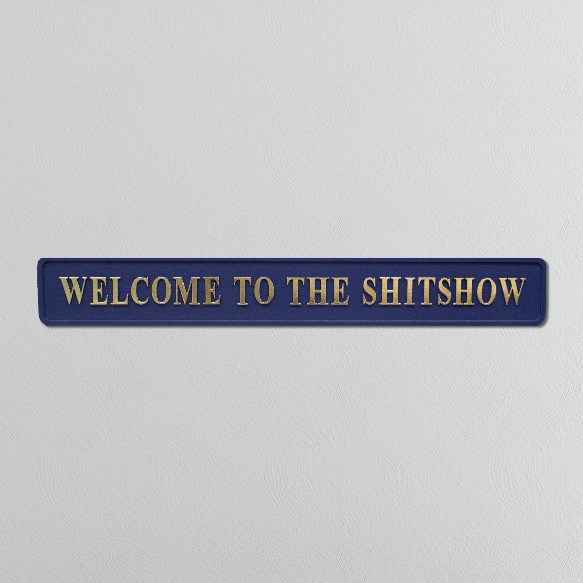 Navy blue sign with gold text 'WELCOME TO THE SHITSHOW' on a light gray background