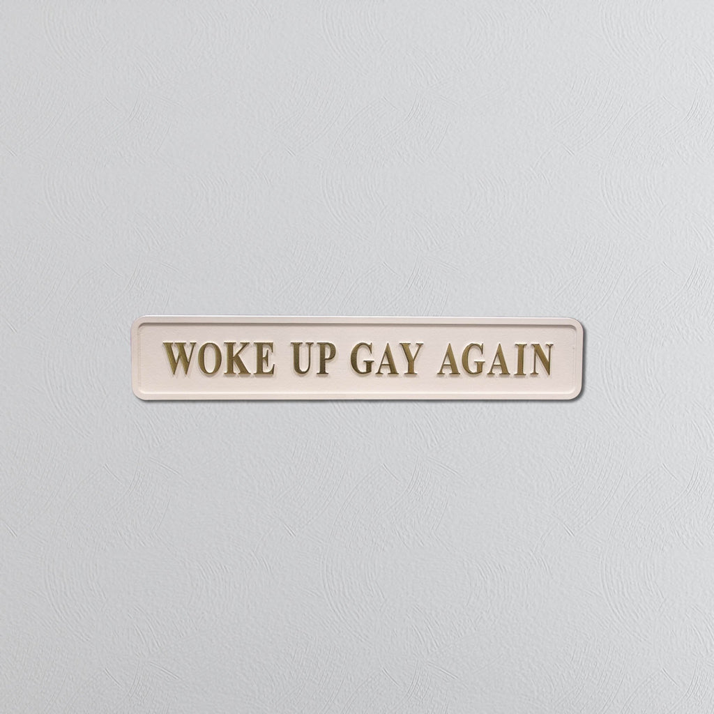 Cream sign with 'WOKE UP GAY AGAIN' in gold text on a light gray background