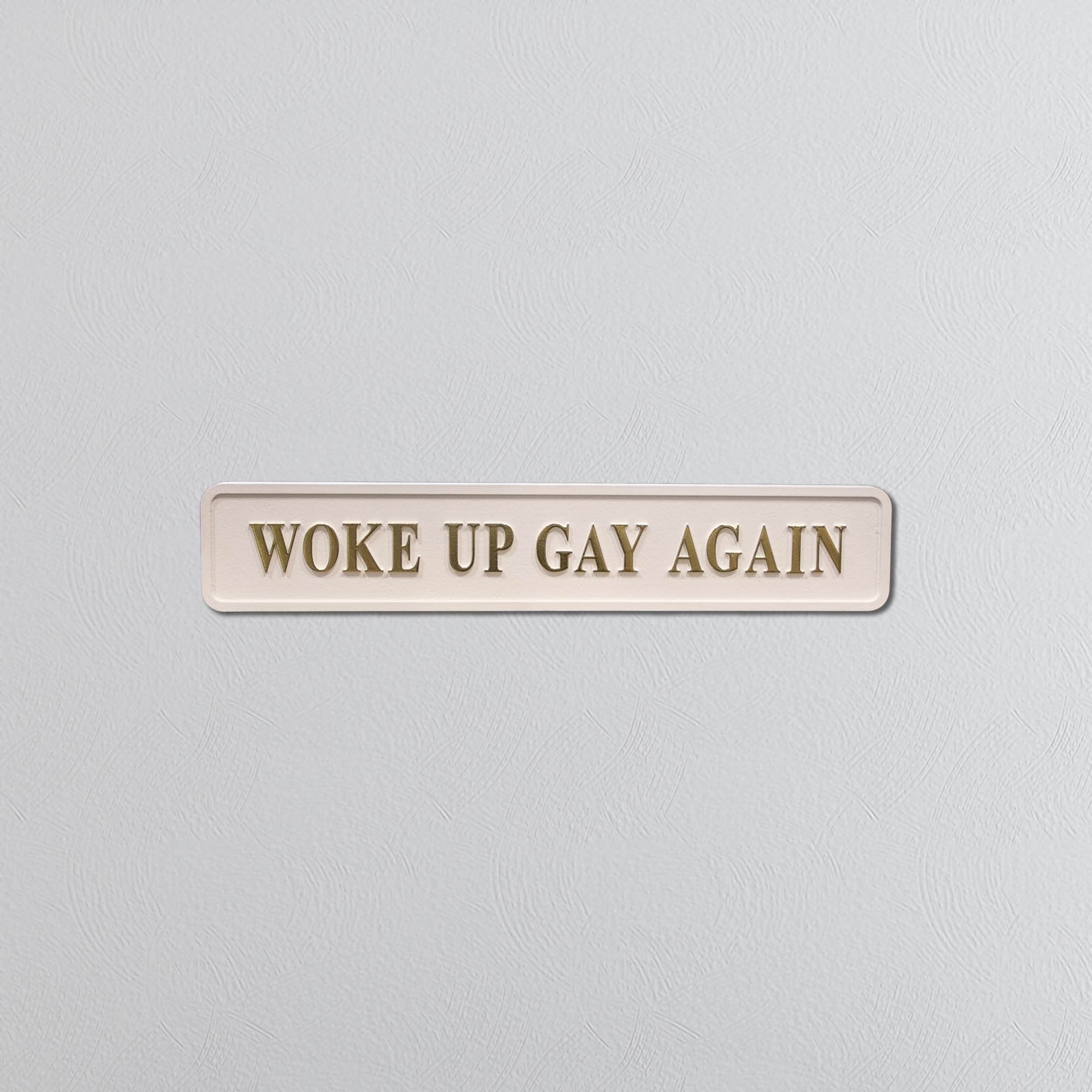 Cream sign with 'WOKE UP GAY AGAIN' in gold text on a light gray background