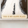 Wooden sign with 'This is my happy place' text in gold in front of an abstract painting