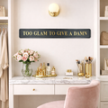Decorative vanity setup with 'Too Glam to Give a Damn' black and gold sign, flowers, and makeup products.