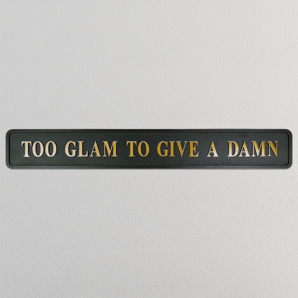 Black sign with gold text 'TOO GLAM TO GIVE A DAMN' on a light gray background