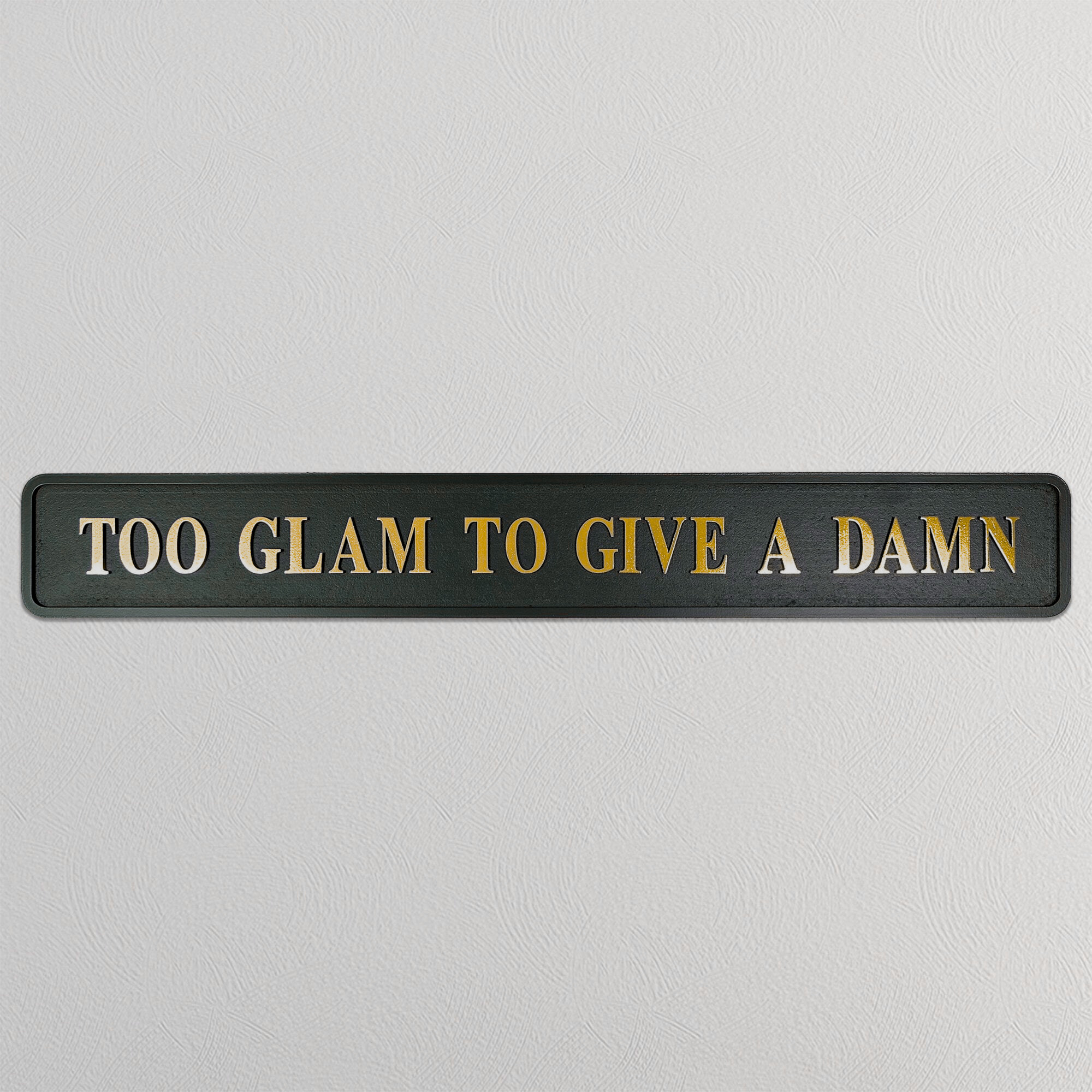 Black sign with gold text 'TOO GLAM TO GIVE A DAMN' on a light gray background