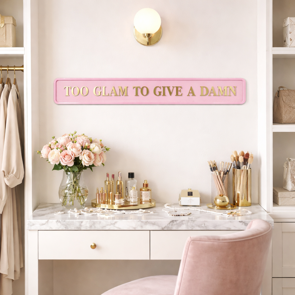 Pink sign with gold text reading 'TOO GLAM TO GIVE A DAMN' on a vanity with flowers and makeup.