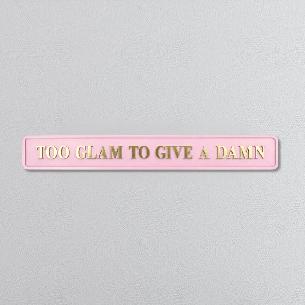 Pink sign with gold text 'TOO GLAM TO GIVE A DAMN' on a light gray background