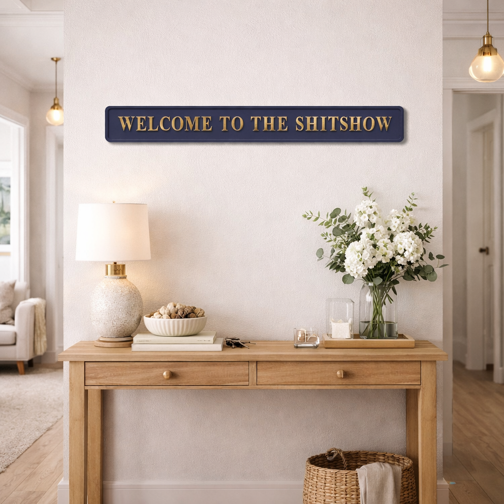 Blue wall sign with 'WELCOME TO THE SHITSHOW' gold text above a wooden console table in a home interior.