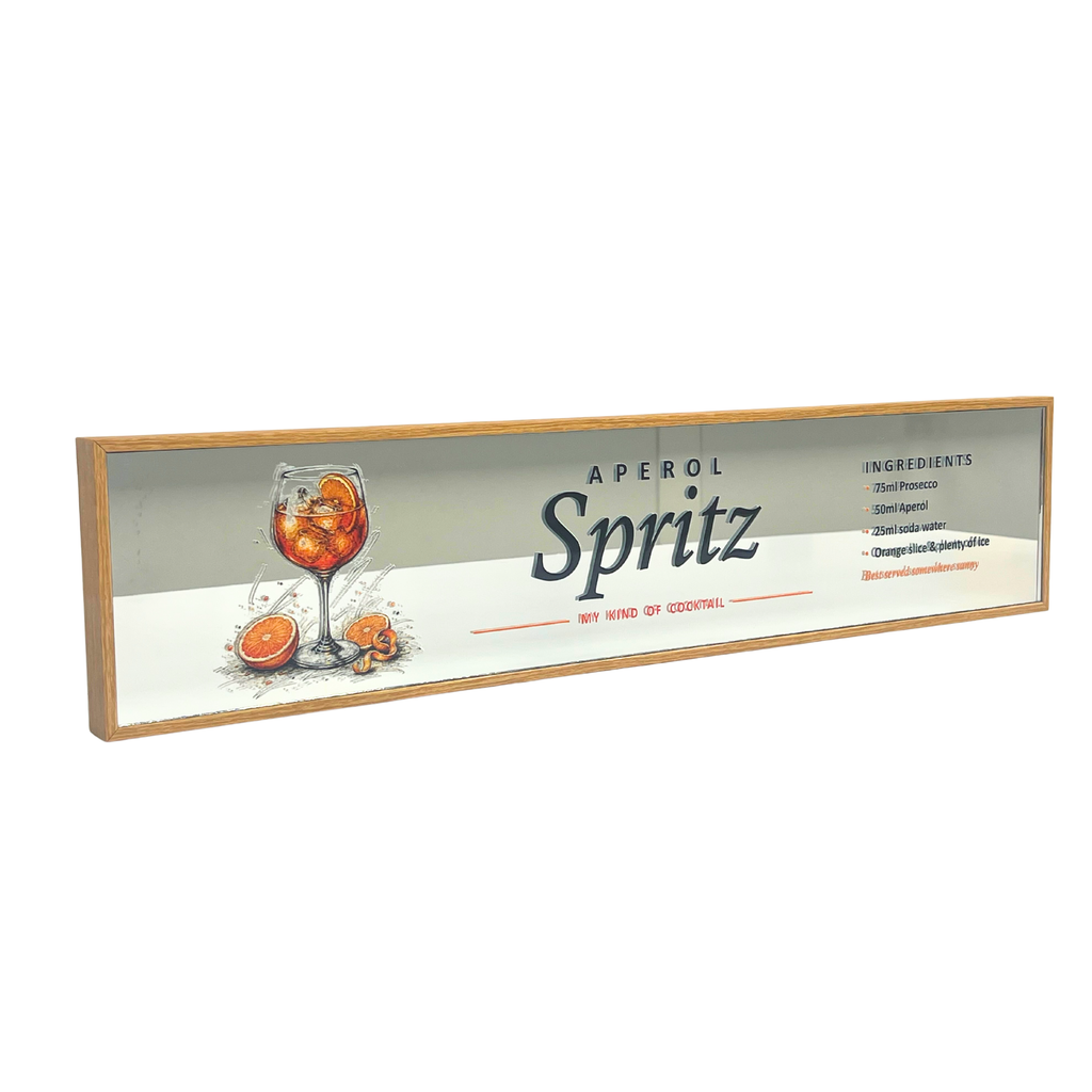 A rectangular framed sign featuring a vintage-style Aperol Spritz cocktail advertisement with a glass of orange-colored drink, orange slices, and printed text on a light background, enclosed in a natural wood frame.
