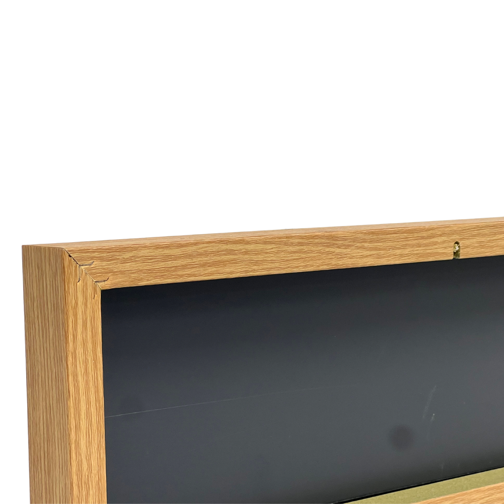 Close-up of the top-left corner of a framed wall art piece with a light oak wood frame and a dark backing board, shown against a plain white background.